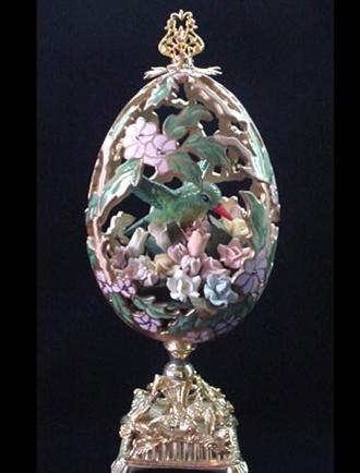 Hand carved w/extreme care and dedication. Top opens showing the hummingbird in his niche full of porcelain roses. Painted in matte satin pink and green with gold enamel details. Accented w/amethyst and AB Austrian Crystals. Gold glazed 18K gold pigments on the inside. Gold plated heavy stand and findings. Extremely delicate. All-around hand carved. Decorated box available for this eggypiece.