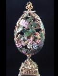 Hummingbird Niche Masterpiece Eggypiece- Hand carved w/extreme care and dedication. Top opens showing the hummingbird in his niche full of porcelain roses. Painted in matte satin pink and green with gold enamel details. Accented w/amethyst and AB Austrian Crystals. Gold glazed 18K gold pigments on the inside. Gold plated heavy stand and findings. Extremely delicate. All-around hand carved. Decorated box available for this eggypiece.